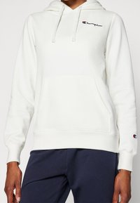 Bílá mikina s kapucí má klokaní kapsu, žebrované manžety a lem, a na levé hrudi je vyšitý logo Champion.