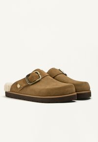 Hnedé semišové klompy s mäkkou kožušinovou podšívkou, vybavené širokým remienkom a kovovou prackou. Hrubá gumová podrážka, hladká textúra a zaoblený tvar.