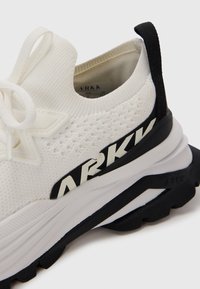 Zapatilla de punto blanca con un upper texturizado, contrafuerte negro y logo de ARKK en negro en el lateral. Presenta una suela duradera y cierre de cordones.