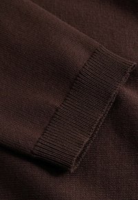 Tmavohnedá pletená látka so zloženým manžetovým rukávom s rebrovanou textúrou a jemnými detailmi šitia.