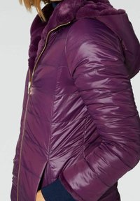 Chaqueta acolchada morada con capucha forrada de piel sintética y cierre de cremallera. La tela es suave y acolchada, con puños acanalados para mayor calidez.