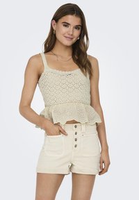 Šviesiai beige megztas viršus su peplum apdaila ir banguotais kraštais, derinamas su aukštu liemeniu grietinėlės spalvos šortais, turinčiais mygtukų užsegimą ir kišenes.