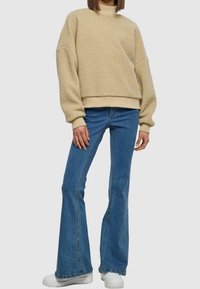Beige Fleece-Pullover mit rundem Halsausschnitt und überschnittenen Schultern, kombiniert mit hellblauen Jeans im Schlag und weißen Turnschuhen.