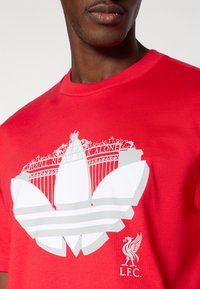 Κόκκινο βαμβακερό T-shirt με μεγάλο λευκό γραφικό ενός πουλιού και το κείμενο «YOU'LL NEVER WALK ALONE» από πάνω, με ένα λογότυπο στη γωνία.