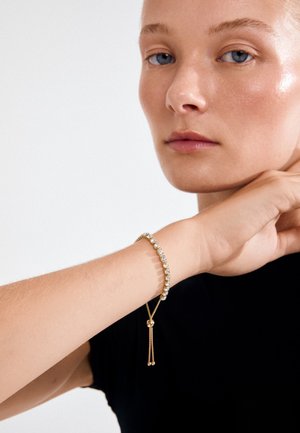 Gold bracelet with clear gemstones, featuring a delicate chain and a sliding clasp. Worn on a wrist above a black shirt.