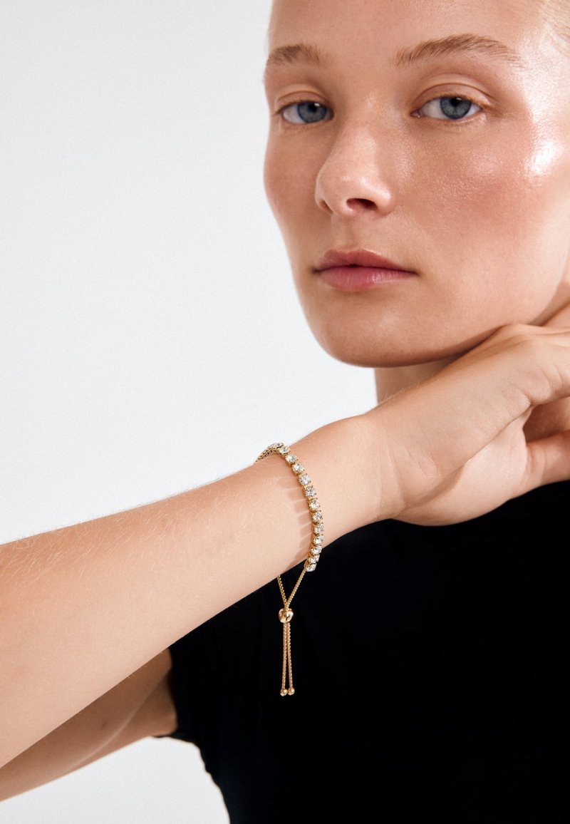 Zelta rokassprādze ar caurspīdīgiem dārgakmeņiem, kurai ir smalka ķēde un slīdoša aizdare. Nēsāta uz rokas virs melnas kreklu.