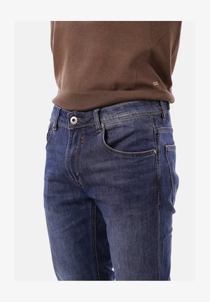 Gros plan sur la taille portant un jean en denim bleu avec des taches et un pull en tricot marron rentré à l'intérieur.