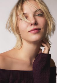 Off-shoulder, ribgebreid truitje in diep paars; het haar van het model is lichtblond, met zachte golven, en ze houdt haar hals vast.