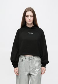 Sweatshirt preta cropped com capuz ajustável e mangas raglan, com letras brancas "PINKO" na frente; combinada com calças prateadas brilhantes.