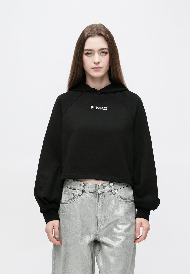 Sweatshirt preta cropped com capuz ajustável e mangas raglan, com letras brancas "PINKO" na frente; combinada com calças prateadas brilhantes.