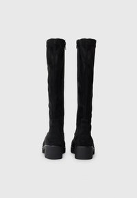 Paire de bottes hautes en daim noir avec talons larges et bas, et fermetures éclair sur le côté, vues de dos sur un fond clair uni.