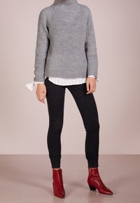 Person, der einen grauen gerippten Pullover über einem weißen gestreiften Hemd, schwarze enge Jeans und rote Absatz-Ankle-Boots trägt, vor einem neutralen Hintergrund.