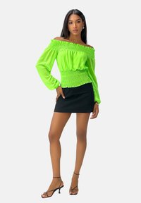 Groene off-the-shoulder blouse met gerimpelde taille, lange mouwen en een soepele textuur, gecombineerd met een zwarte mini-jurk en sleehakken.
