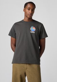 Grey cotton t-shirt with short sleeves, featuring a colorful graphic logo on the chest. Model wearing beige trousers.