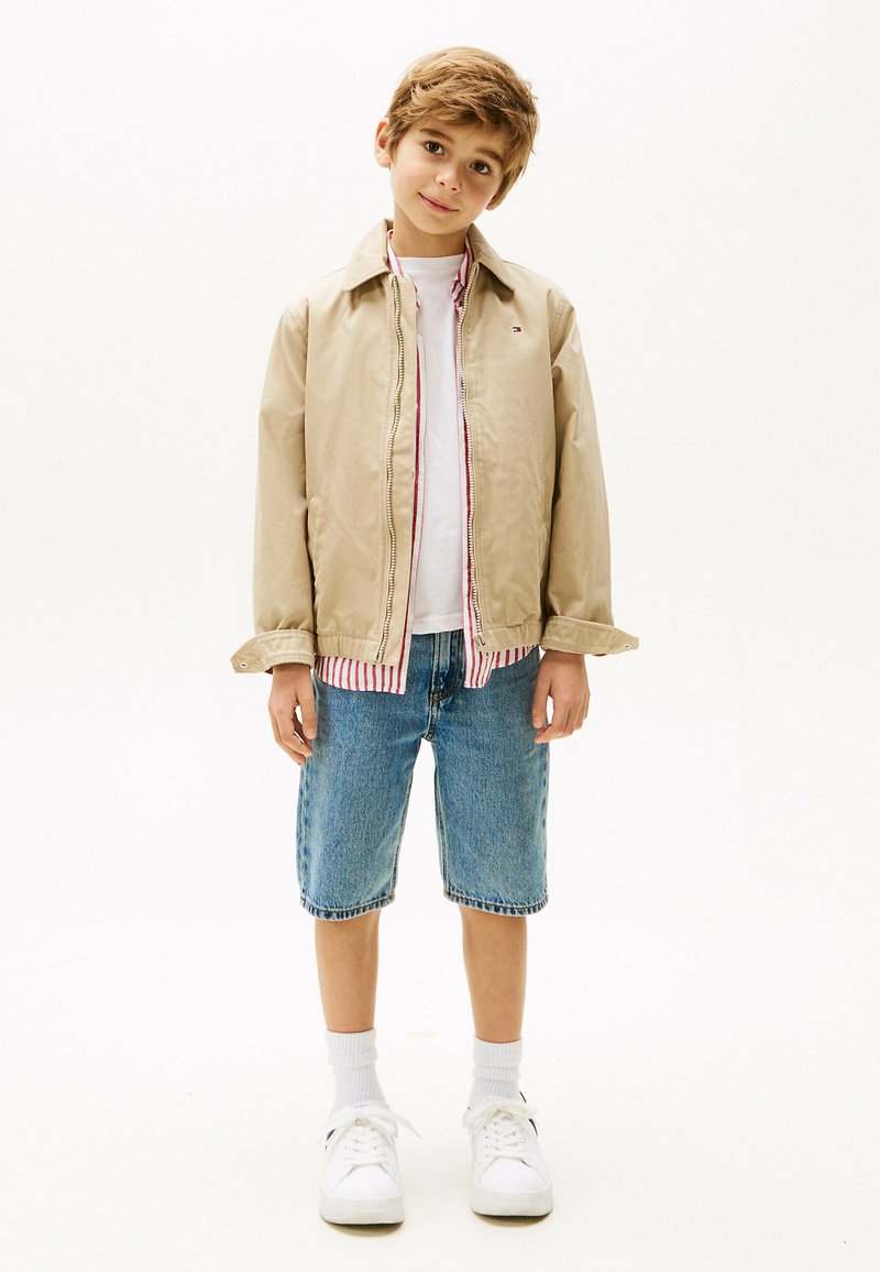 Veste beige avec fermeture éclair, doublure intérieure à rayures rouges, t-shirt blanc, shorts en denim bleu, chaussettes blanches et baskets blanches.