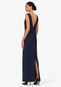 Navy blauwe mouwloze jurk met een diepe V-rug, zijsplit en een slanke silhouet, gemaakt van gladde stof. Draagt bij zwarte hakken.