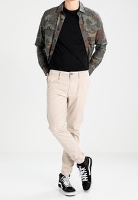 Chemise boutonnée à motif camouflage, t-shirt noir à col rond, pantalon beige slim, baskets noir et blanc. Fond uni.