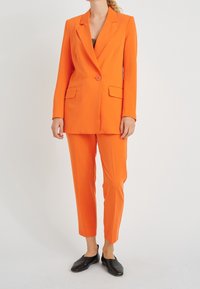 Oranžový oblek s ušitým blejzrem s klopami, dvěma předními kapsami a odpovídajícími zúženými kalhotami. Nosený s černými slip-on botami.