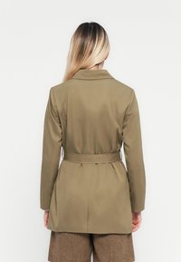 Kobieta z długimi blond włosami, ubrana w oliwkową marynarkę z paskiem i brązowe szorty w jodełkę, stojąca na tle jednolitego białego tła.