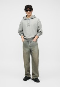 Νεαρός με σγουρά μαλλιά φοράει μαύρα γυαλιά ηλίου, γκρι φούτερ με κουκούλα και μικρό λογότυπο στο μπροστινό μέρος, φαρδιά ξεβαμμένα πράσινα τζιν και μαύρα αθλητικά παπούτσια, με τα χέρια στις τσέπες.