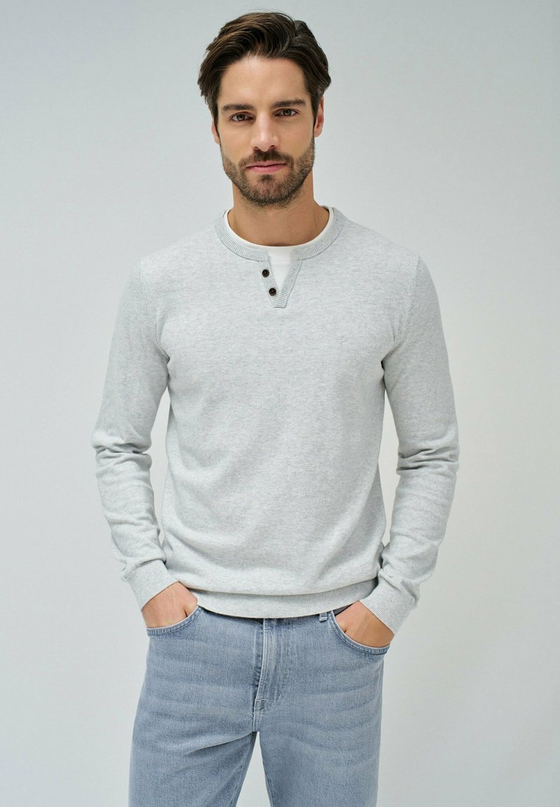 Graues Langarmshirt mit rundem Ausschnitt, das über einen Knopf-Detail am Kragen verfügt. Getragen mit hellblauen Jeans, Hände in den Taschen.