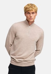 Beige halvdraget tröja med texturerad stickning, ribbad krage och muddar. Buren över svarta byxor, med händerna knäppta bakom ryggen.