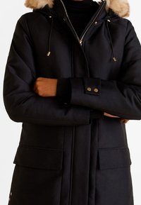 Zwarte parka jas met een capuchon gevoerd met bont, gouden accenten van hardware, een ritssluiting en voorzakken, gemaakt van een gladde, gestructureerde stof.