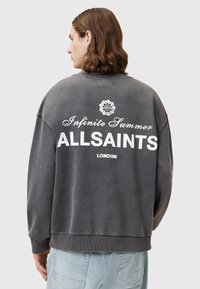 Szara bluza z oversize'owym krojem, okrągłym dekoltem i prążkowanymi mankietami. Na plecach znajduje się biały nadrukowany tekst: "Nieskończone Lato ALLSAINTS LONDYN".