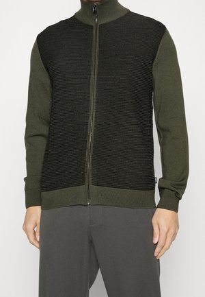 Mann trägt einen olivgrünen Reißverschluss-Pullover mit strukturiertem schwarzem Strickvorderteil und passenden olivgrünen Ärmeln und Kragen.