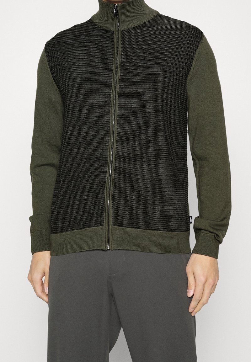 Homme portant un pull zippé vert olive avec un panneau avant en tricot noir texturé et des manches et un col assortis verts olive.