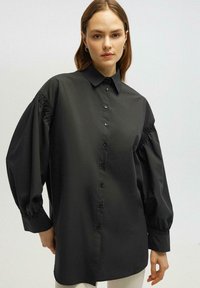 Mustamärkidega särk klassikalise kraega, puffitud pikkade varrukatega ja elastsete mansettidega. Sile tekstuur, vabalt istuv ja sirge alläär.