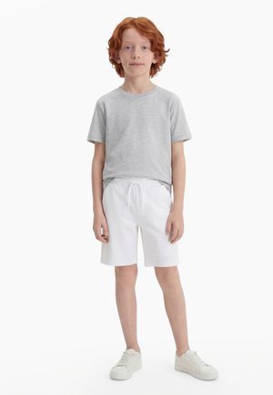 Garçon aux cheveux roux debout, portant une chemise à manches courtes gris clair, un short blanc avec cordon de serrage et des baskets blanches sur fond blanc.