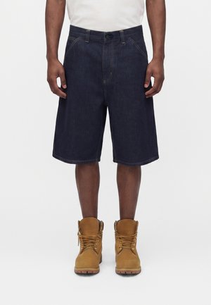 Mand iført mørkeblå denimshorts til knæet og brune snørestøvler, står mod en ensfarvet hvid baggrund.