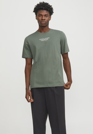 Πράσινη βαμβακερή μπλούζα με κοντά μανίκια, που φέρει τυπωμένο λογότυπο "JACK & JONES PREMIUM" σε λευκό. Φορεμένη με μαύρο παντελόνι.