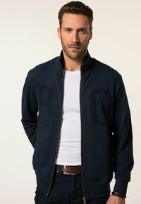 Sudadera azul marino con cremallera y cuello alto, con números bordados en relieve "8" y "0" en el frente, combinada con una camiseta blanca.