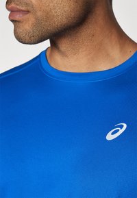 Sinine sportlik särk, lühikeste käistega, valmistatud tekstureeritud kangast, millel on vasakul rinnal väike valge logo. Ümar kaeluse disain.