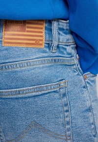 Vaaleansiniset farkut, joissa on nahkapaikka, jossa on lippu design. Farkuissa on kontrastivärinen oranssi tikkaus ja klassinen viiden taskun tyyli.