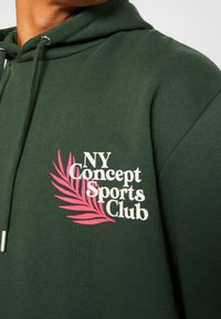 Sudadera verde oscuro con bolsillo canguro, que presenta un gráfico blanco y rosa con el logo "NY Concept Sports Club" y un diseño de hoja en el pecho.