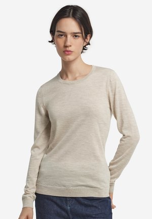 Ung kvinde med kort mørkt hår iført en beige langærmet sweater og mørkeblå jeans, stående med den ene hånd på hoften.