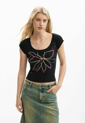 Femme aux longs cheveux blonds portant un haut noir sans manches avec une grande fleur brodée et une jupe en jean bleu délavé.