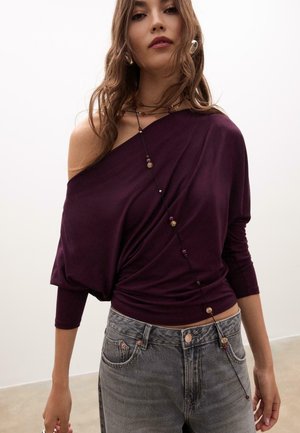 Lila schulterfreies Oberteil aus weichem Stoff mit drapiertem Design, kombiniert mit hoch-taillierten grauen Jeans. Perlenketten betonen den Ausschnitt.