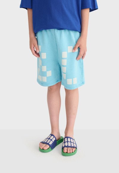 Enfant portant un short bleu clair avec des carrés blancs, une chemise bleue et des sandales à glissière vertes avec des brides bleues et un texte blanc.