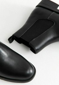 Botas de tobillo negras de cuero liso, con paneles laterales elásticos, punteras redondeadas y suelas texturizadas. Diseño minimalista con acabado mate.