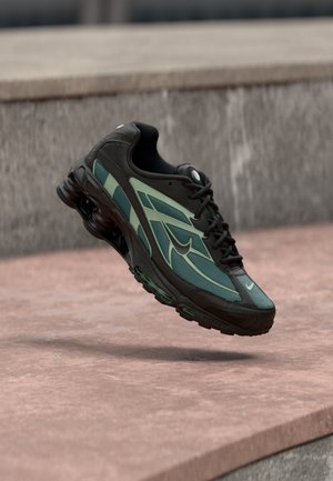 Zapato deportivo para hombre en negro con detalles verdes, que presenta una parte superior de malla, superposiciones texturizadas y un diseño distintivo con talón acolchado.