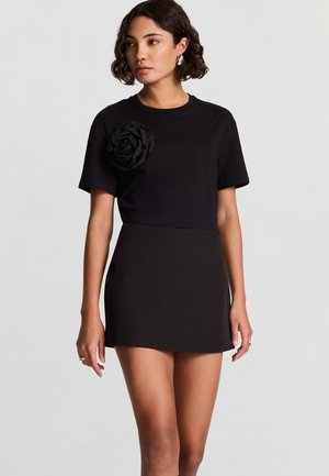 Top nero a maniche corte con scollo rotondo e una rosa di tessuto nero tridimensionale sulla spalla sinistra. Abbinato a una minigonna nera.