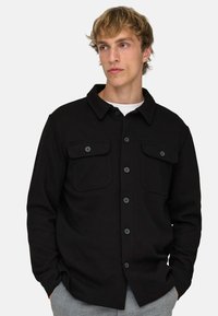 Junger Mann mit blondem Haar, trägt eine schwarze Hemdjacke mit Klappentaschen, ein weißes Unterhemd und graue Hose, Hände in den Taschen.