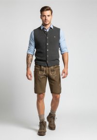 Gilet grigio di lana sopra una camicia azzurro chiaro, abbinato a pantaloni corti marroni al ginocchio con ricami decorativi. Stivali da trekking marroni completano l'outfit.