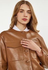 Braune Lederhemdjacke mit glänzendem Finish, ausgestattet mit einem weißen Kragen, zwei Brusttaschen und Druckknopfverschlüssen.