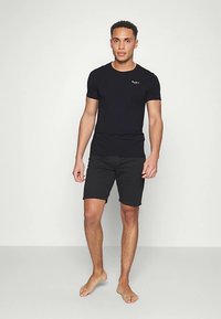 Čierne tričko s krátkym rukávom a okrúhlym výstrihom s jemným logom na hrudi, v kombinácii s čiernymi šortkami, ktoré má na sebe model stojaci naboso.