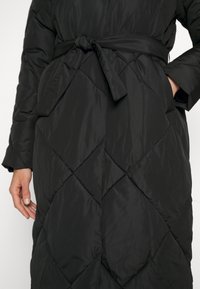 Lange zwarte pufferjas met een ruitpatroon, met een ceintuur in de taille. Het materiaal lijkt glad met een lichte, gewatteerde textuur.